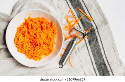 Salad Of Fresh Shredded Carrots In A White Bowl Next To A Carrot Shredder On A Light Background. Korean Carrot. Top View. Healthy Food Concept.