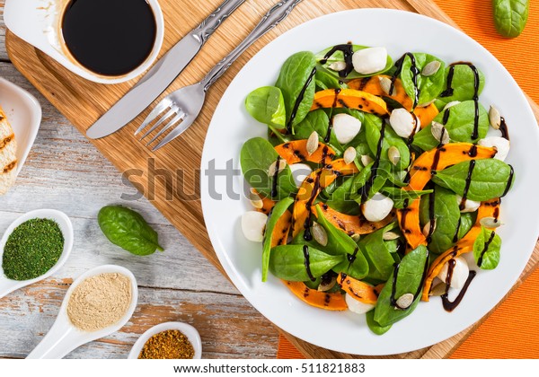 salad of grilled pumpkin, mini mozzarella and\
spinach, seasoned with squash seeds on white plate on chopping\
board with fork and knife, spices and balsamic vinaigrette as \
dressing, view from above