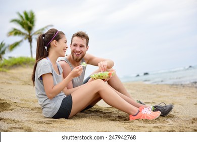 Salad - Healthy Fitness Woman And Man Couple Laughing Eating Food Lunch Sitting On Beach After Workout. Mixed Race Asian Caucasian Female Model And Male Models In Sportswear.