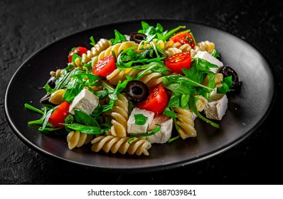Salat mit Nudeln, Arugula, Tomate, Käse und Oliven auf schwarzem Teller auf schwarz-grauem schwarzem Hintergrund