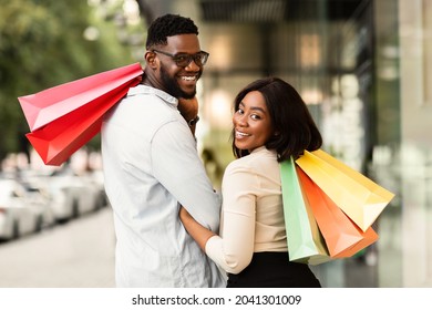 Sale And Consumerism Concept. Portrait Of Happy African American Couple Holding Shopping Bags. Smiling Black Man And Woman Carrying Purchases, Turning Back And Looking At Camera Over The Shoulder