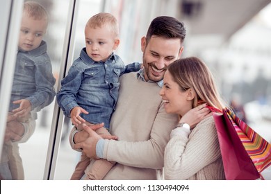 Sale, Consumerism And Family Concept - Happy Young Family With Shopping Bags And Their Son, Looking At Shop Window In City.