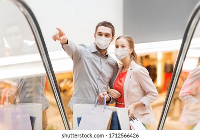 sale, consumerism and pandemic concept - happy young couple wearing face protective medical mask for protection from virus with shopping bags rising on escalator and pointing finger in mall - Powered by Shutterstock