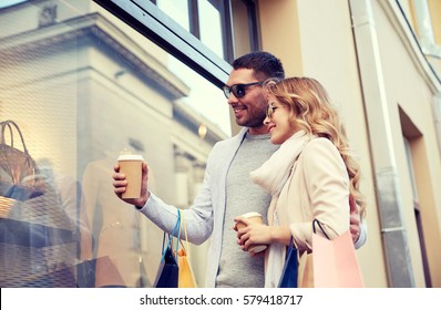 Sale, Consumerism And People Concept - Happy Couple With Shopping Bags And Coffee Paper Cups Looking At Shop Window In City