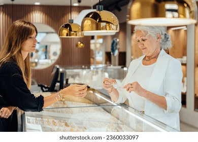 Sale, consumerism, shopping and people concept. Happy grey haired elderly woman buying some jewelry. Blond woman owner, saleswoman of jewelry shop presenting for senior female consumer golden earrings - Powered by Shutterstock