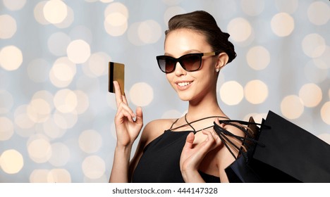 Sale, Finances, Fashion, People And Luxury Concept - Happy Beautiful Young Woman In Black Sunglasses With Credit Card And Shopping Bags Over Holidays Lights Background