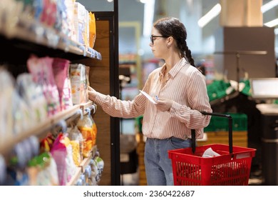 Sale in grocery store. Side view of young caucasian woman wearing eyeglasses holds cart and take food from upper shelf. Concept of shopping in supermarket and consumerism. - Powered by Shutterstock