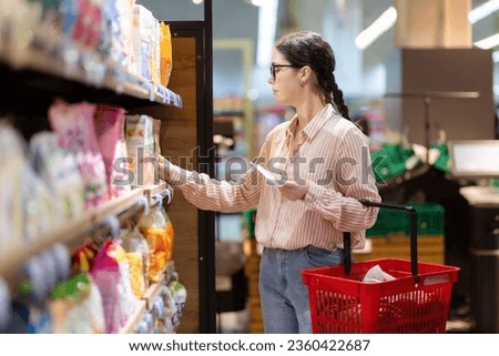 Sale in grocery store. Side view of young caucasian woman wearing eyeglasses holds cart and take food from upper shelf. Concept of shopping in supermarket and consumerism.