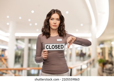 Sale, Lockdown And Business Concept - Female Shop Assistant With Closed Sign And Name Tag Showing Thumbs Down Over Mall Background