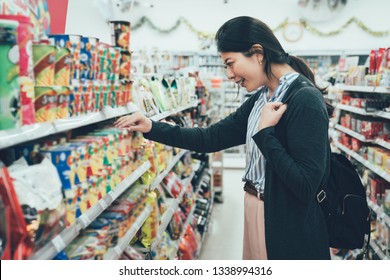 sale shopping consumerism people concept. asian woman backpacker with curious face picking snack cookies choose at grocery supermarket. lady buying products food instant noodles in convenience store. - Shutterstock ID 1338994316