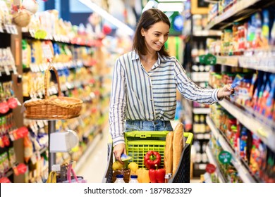 Sale, Shopping, Consumerism And People Concept. Smiling young woman standing with trolley basket full of eco bio products, walking near shelves at grocery store, choosing healthy food in market - Shutterstock ID 1879823329