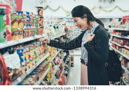sale shopping consumerism people concept. asian woman backpacker with curious face picking snack cookies choose at grocery supermarket. lady buying products food instant noodles in convenience store.