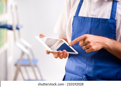 Sales Clerk Wearing Apron Using A Digital Tablet With Store Shelves On Background.