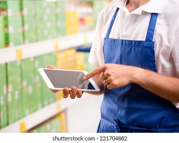 Sales Clerk Wearing Apron Using A Digital Tablet With Store Shelves On Background.