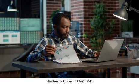 Sales consultant analyzing financial charts on papers and laptop, using statistics to plan new stock market profit strategy. Working at night with hedge fund exchange, forex trade and numbers.