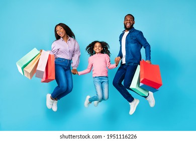 Sales And Discounts. Full Body Length Shot Of Excited African American Family Of Three People Jumping, Holding Hands And Shopping Bags With New Clothes, Rejoice Purchase, Blue Studio Wall