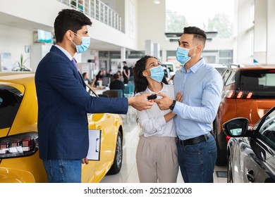 Sales Manager Giving Happy Middle-eastern Young Family Key From Brand New Auto, Wearing Protective Face Masks, Side View. Arabic Couple Buying Car At Luxury Delarship Salon. Car Purchasing Concept