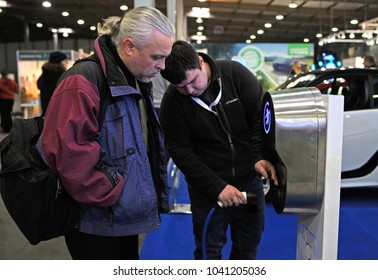 Sales Manager Holds Electric Charger Pluged In A Charging Station Explaining To A Customer How To Charge An Electric Car. Exhibition PLUG-IN UKRAINE 2018. March 2, 2018. Kiev Expo Center. Kiev,Ukraine