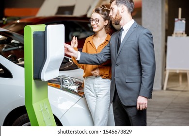 Sales Manager Showing Car Charging Station To A Young Woman In The Showroom With Electric Cars At The Car Dealership