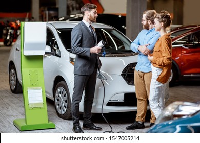 Sales Manager Showing Car Charging Station To A Young Couple, Selling Electric Cars In The Showroom. Concept Of Buying Eco-friendly Car For Family