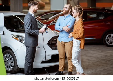 Sales Manager Showing Car Charging Station To A Young Couple, Selling Electric Cars In The Showroom. Concept Of Buying Eco-friendly Car For Family