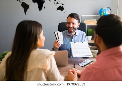 Sales Representative Giving Their Plane Tickets And Travel Itinerary To An Hispanic Couple. Young Man And Woman Booking A Cruise For Their Honeymoon