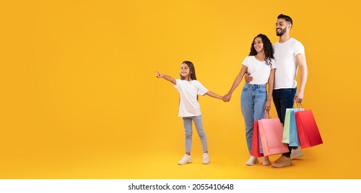Sales Season. Arabic Family On Shopping Holding Colorful Paper Shopper Bags Pointing Finger Aside Posing Standing On Yellow Studio Background. Panorama With Free Space For Text