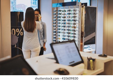 Saleslady Assisting A Customer In A Store Trying On Eyeglasses With A View Of A Computer Terminal In The Foreground