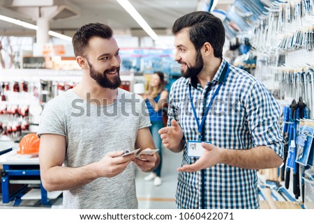 Salesman in checkered shirt is showing bearded client selection of equipment in power tools store.