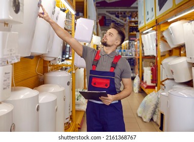 Salesman Checking One Of The Store Aisles. Young Man In Work Uniform Holding Clipboard, Walking Along The Aisle And Looking At Wide Range Of Modern Electric Water Heaters. Retail Business Concept