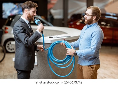 Salesman Giving Cable For Electric Car Charging To A Client In The Car Dealership. Man Buying Electric Car