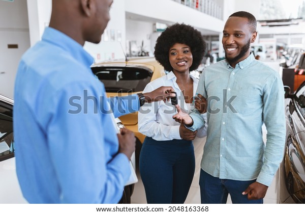 Salesman giving car key to\
young black couple at auto dealership. African American clients\
buying or renting automobile at showroom. Vehicle local\
distribution concept