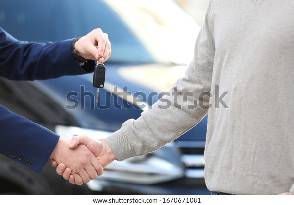 Salesman giving key\
to customer while shaking hands in modern auto dealership, closeup.\
Buying new car