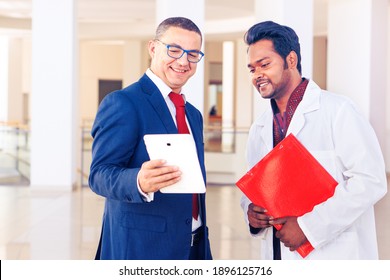 Salesman and indian doctor meet at hospital hall - Businessman and medical staff talk in the clinic lobby looking at tablet device - Concept of healthcare business , work and technology - Image 