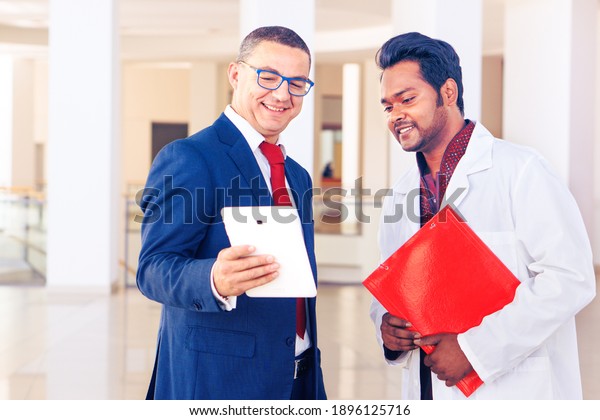 Salesman and indian doctor meet at hospital hall -\
Businessman and medical staff talk in the clinic lobby looking at\
tablet device - Concept of healthcare business , work and\
technology - Image 