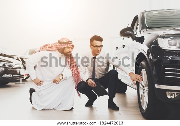 Salesman points to\
wheels of new car and smiles. Arab man bent to check wheels in a\
car with a young seller.\
