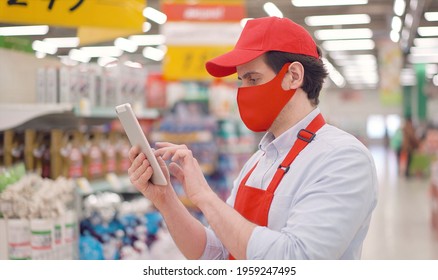 Salesman in red face mask and uniform standing in supermarket and typing on tablet computer. Deliveryman working in delivery service during epidemic covid-19, online food shopping - Powered by Shutterstock