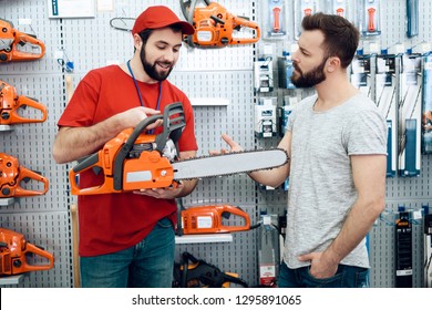 Salesman In Red Shirt And Baseball Cap Is Showing Bearded Client New Chainsaw In Power Tools Store.