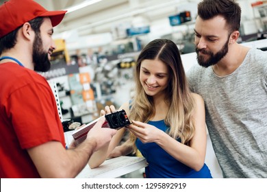 Salesman In Red Shirt And Baseball Cap Is Showing Couple Of Clients Power Tool In Power Tools Store.