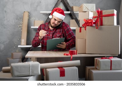 A Salesman In A Santa Claus Hat Takes An Order By Cell Phone While Sitting In An Office With Boxes Of Gifts And Surprises For Christmas For Office Workers. Shopping And Delivery. Small Business.