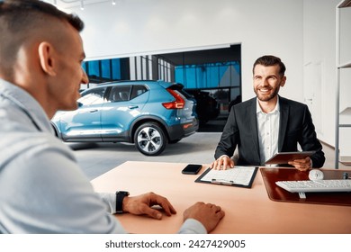 A salesman in a suit sitting at a table in a car showroom with a client and drawing up documents using a tablet and computer. Buying, renting and insuring a car. - Powered by Shutterstock