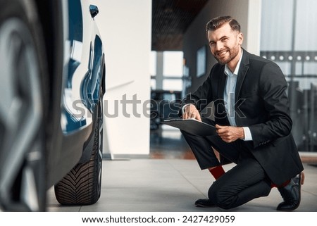 A salesman in a suit with a tablet stands in a car showroom and demonstrates the car to buyers, pointing to the wheels. Buying, renting and insuring a car.