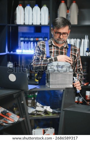 Salesman using barcode scanner on car battery in auto parts store, surrounded by shelves of products and using a laptop