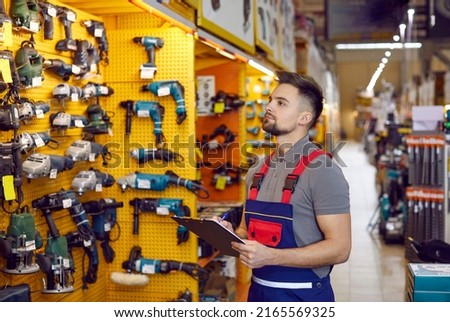 Salesman writing on a clipboard while checking construction and carpentry tools on display. Young man who works at a DIY store or hypermarket looking at various tools on shelves in one of departments