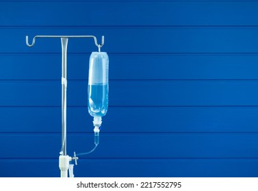 Saline Bag Hanging On Poles Stand Isolated On Blue Wood Plank Wall Background With Copy Space. Saline Solution IV Drip Fluid Bottle For Infusion For Patient In Ward Hospital.