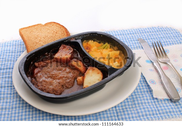 Salisbury steak and potatoes TV\
dinner in plastic dish on white plate against blue gingham place\
mat.