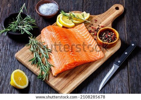 salmon fillet with peppercorn, rosemary and lemon on wooden cutting board on dark wood table with knife, horizontal view from above