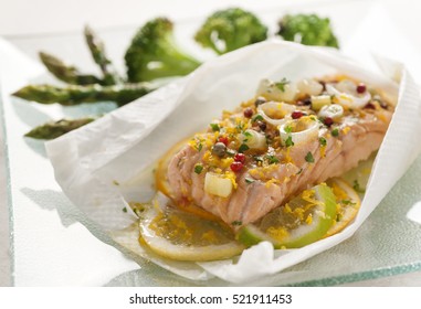 salmon loin wrapped in kitchen paper with citric fruits, orange, lime, lemon, seasoned with onion, pepper, parsley served with broccoli and green asparagus on a glass tray