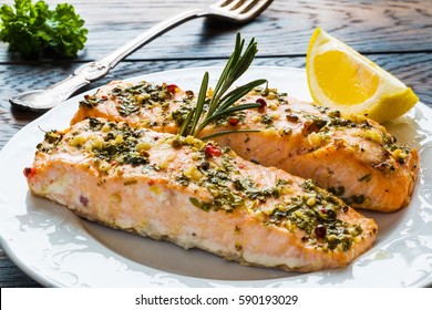 Salmon roasted in an oven with a butter, parsley and garlic. Portion of cooked fish and fresh lemon on a white plate on the wooden table