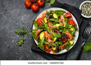 Salmon salad with green leaves, avocado and tomato on black stone background. Top view with copy space.